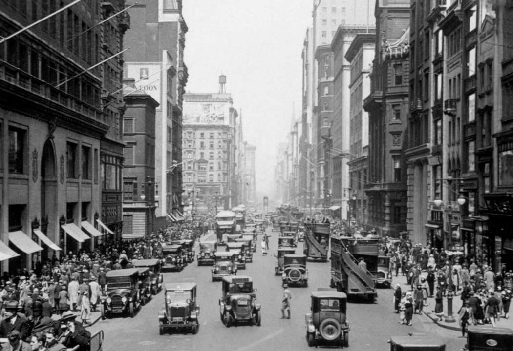 Historical black and white image of a bustling city street, populated by pedestrians and cars.